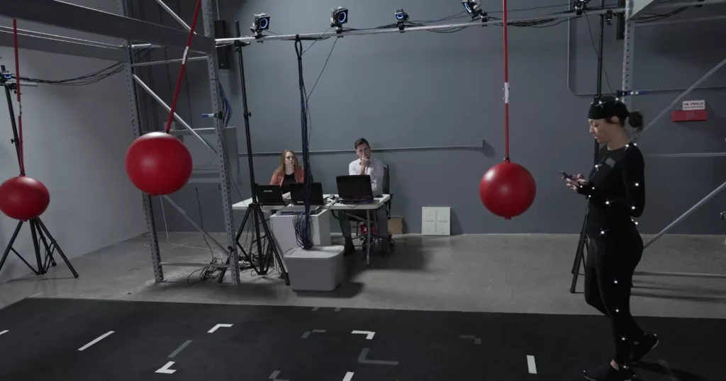 A performer wearing motion-capture markers walks through a studio while technicians monitor the data for game animation.